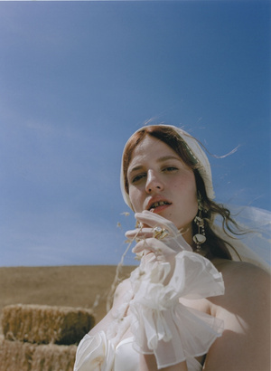 Woman in white dress and headscarf posing outdoors in rural landscape with blue sky and hay bales, fashion photography, portrait.