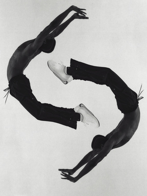 Two shirtless African American adult males pose in an arching backbend, one upside down, against a solid white background, wearing dark pants and white sneakers.