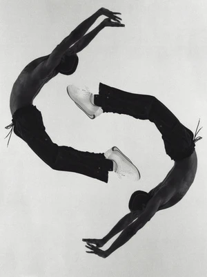 Two shirtless African American adult males pose in an arching backbend, one upside down, against a solid white background, wearing dark pants and white sneakers.