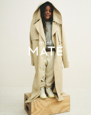 Young child in a tan trench coat and casual clothes stands on a wooden crate in a studio environment, smiling at the camera.