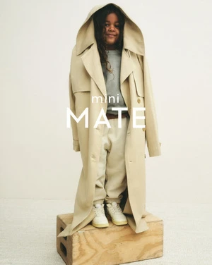 Young child in a tan trench coat and casual clothes stands on a wooden crate in a studio environment, smiling at the camera.