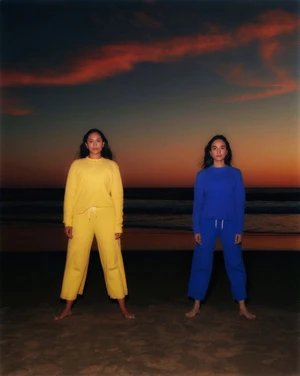 Two women stand on a sandy beach at dusk wearing matching casual outfits in bright yellow and blue, the ocean visible behind them.