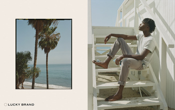 Person sitting on steps near beach with palm trees. Clear sky and ocean visible. Relaxed pose, casual clothing. Lucky Brand logo.