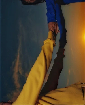 Two people wearing a blue sweatshirt and a yellow sweatshirt holding hands against a warm sunset sky backdrop.