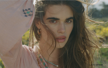 Portrait of a woman with long brown hair, wearing a pink top, outdoors with a field of yellow flowers in the background.