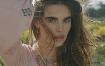 Portrait of a woman with long brown hair, wearing a pink top, outdoors with a field of yellow flowers in the background.