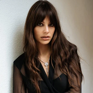 Portrait of a woman with long dark wavy hair, heavy eye makeup, and a diamond pendant necklace wearing a black top.