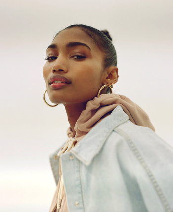 Portrait of a Black woman with gold hoop earrings and a denim jacket against a plain background, looking confidently at the camera.
