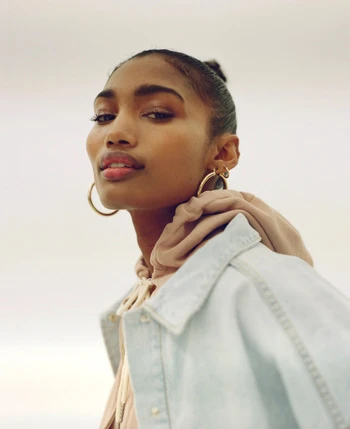 Portrait of a Black woman with gold hoop earrings and a denim jacket against a plain background, looking confidently at the camera.