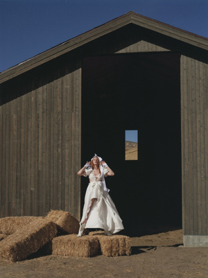 Fashionable woman in white dress and headpiece poses on hay bales in front of barn, bathed in sunlight against blue sky backdrop.