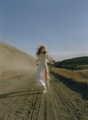A woman in a flowing white dress runs along a dusty road with fields and hills under a bright sky, creating a windswept effect.