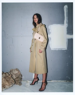 Woman in beige trench coat and black strappy heels standing against a textured grey wall holding a light pink shoulder bag.