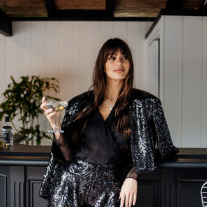 Adult female in sequin shorts and jacket holds a martini in an indoor bar setting with dark wood trim and neutral walls.