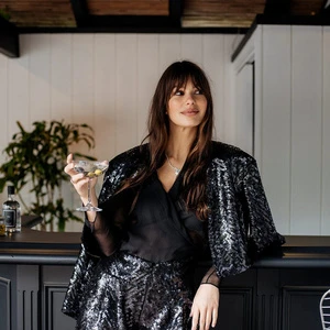 Adult female in sequin shorts and jacket holds a martini in an indoor bar setting with dark wood trim and neutral walls.