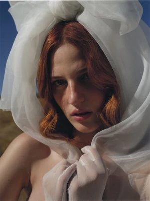 Close-up portrait of a redhead woman with pale skin wearing sheer white headscarf and gloves against a blue sky and neutral ground.