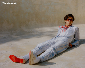 An adult man in a casual suit and red shoes reclines on a concrete surface, bathed in sunlight, shot at eye-level in an outdoor setting.
