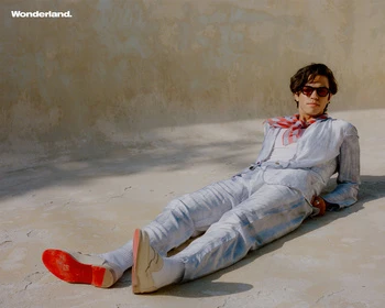 An adult man in a casual suit and red shoes reclines on a concrete surface, bathed in sunlight, shot at eye-level in an outdoor setting.