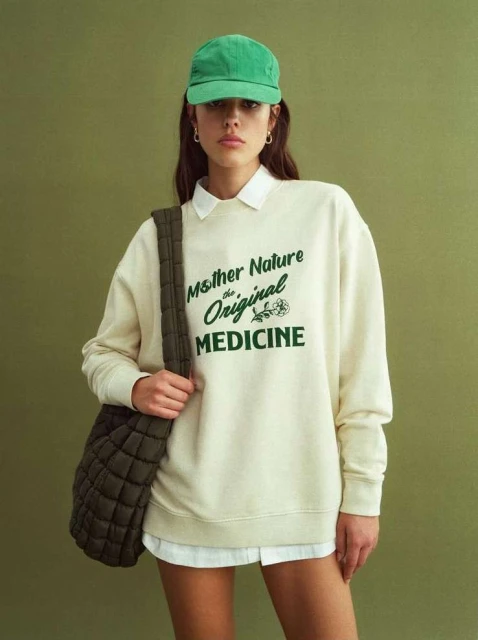 A woman in a sweatshirt and baseball cap poses in front of an olive-green background, carrying a quilted bag over her shoulder.