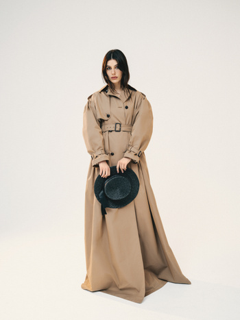 Woman in a long tan trench coat poses in a studio holding a black hat. Fashion editorial image featuring dramatic studio lighting.