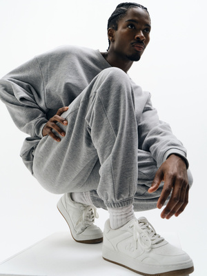 Adult male poses in a gray sweatsuit with white sneakers on a white surface in a studio setting, looking off-camera with a neutral expression.