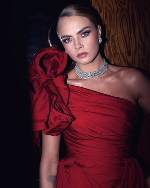 Portrait of Cara Delevingne in a red gown with a large rosette on the shoulder, wearing diamond jewelry, against a dark wood background.
