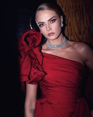 Portrait of Cara Delevingne in a red gown with a large rosette on the shoulder, wearing diamond jewelry, against a dark wood background.
