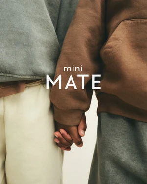 Two children stand shoulder to shoulder in an eye-level medium shot, holding hands, wearing matching loungewear, displaying the mini MATE logo.
