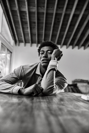 A portrait photograph shows a Black male model against a dark grey backdrop in a photo studio, lit with professional lighting equipment.