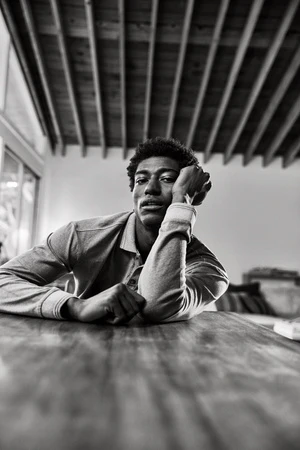 A portrait photograph shows a Black male model against a dark grey backdrop in a photo studio, lit with professional lighting equipment.