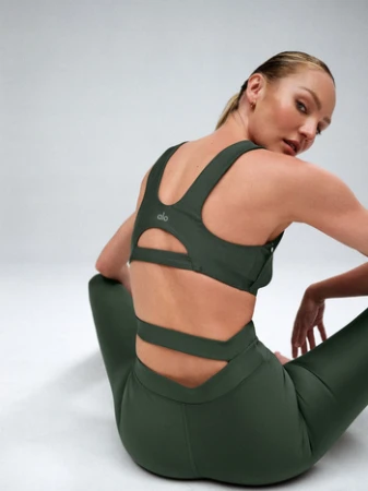 Model seated on white seamless background wearing dark green sports bra and leggings looking back over shoulder