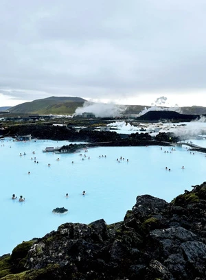People relaxing in the blue waters of a geothermal spa with distant land and steam under an overcast sky, with black rock in the foreground.
