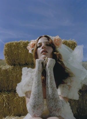 A young woman in a vintage white gown with lace gloves poses outdoors against hay bales under a bright blue sky.