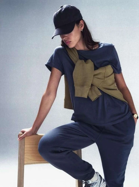 Woman in casual blue shirt and sweatpants posing with wooden chair in studio, baseball cap, sweater, athletic shoes, and neutral background.