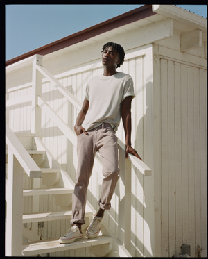 Full-body shot of a Black man in a t-shirt and tan pants posing on white stairs outside on a sunny day with blue sky background.
