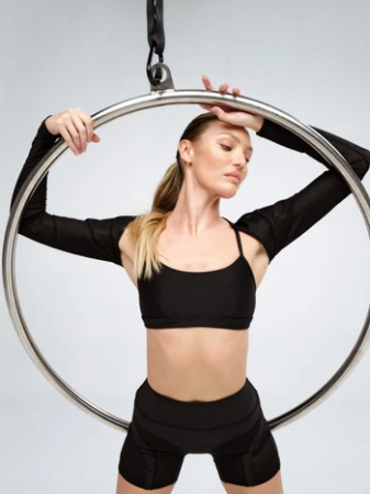 Model posing inside a large suspended metal hoop wearing black activewear against a plain grey studio background.