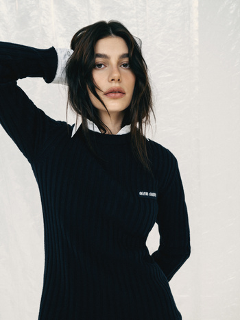 A female fashion model with dark hair wearing a black, long-sleeved knit dress and white collared shirt poses in a studio environment.