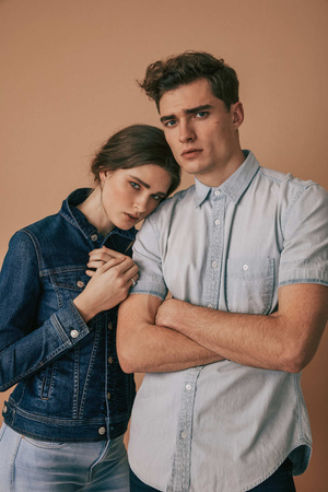 Portrait of a Caucasian couple in light-wash denim outfits. The woman rests her head on the man's shoulder against a plain background.