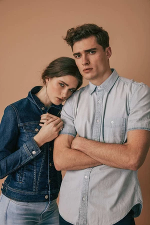 A man in a light blue short-sleeve shirt with arms crossed alongside a woman in a dark denim jacket leaning on him.