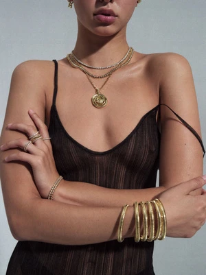 A woman with layered gold necklaces, bracelets, rings, and a sheer black dress is posing in a studio with a neutral background.