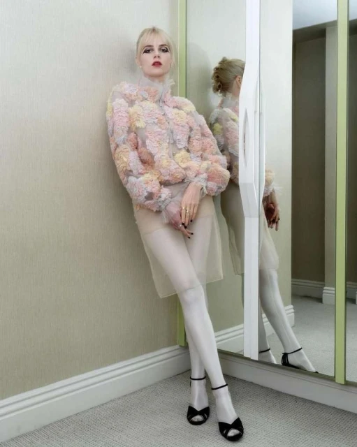 Blonde woman posing against a wall in a floral jacket, sheer skirt, stockings, and sandals in an indoor setting with a mirror.