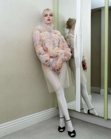 Blonde woman posing against a wall in a floral jacket, sheer skirt, stockings, and sandals in an indoor setting with a mirror.