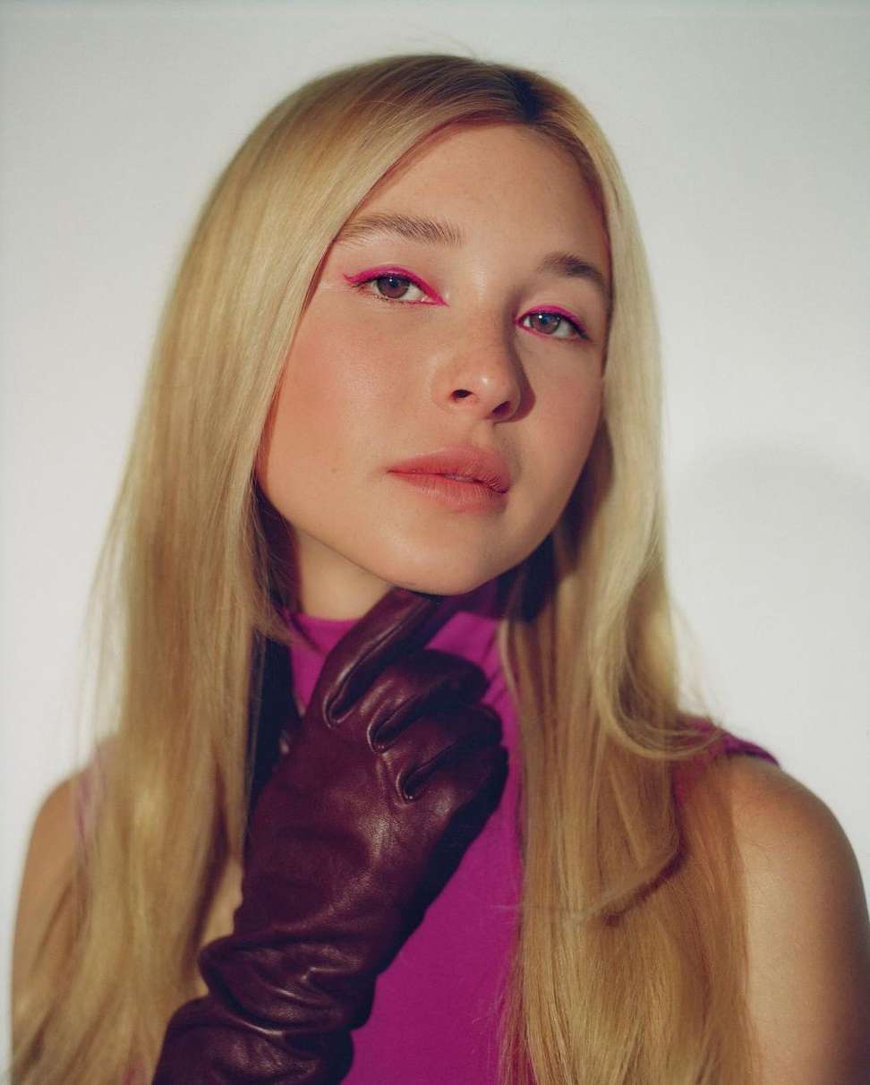 Close-up portrait of a young woman with blonde hair wearing pink eyeliner and maroon leather gloves against a white backdrop.
