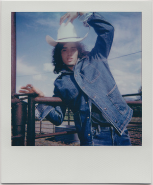 A person in a denim jacket and cowboy hat poses against a rural landscape under a blue sky in a vintage-style, square Polaroid photo.