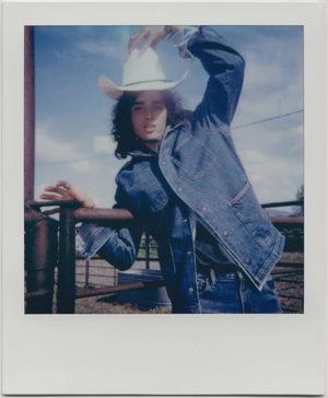 A person in a denim jacket and cowboy hat poses against a rural landscape under a blue sky in a vintage-style, square Polaroid photo.
