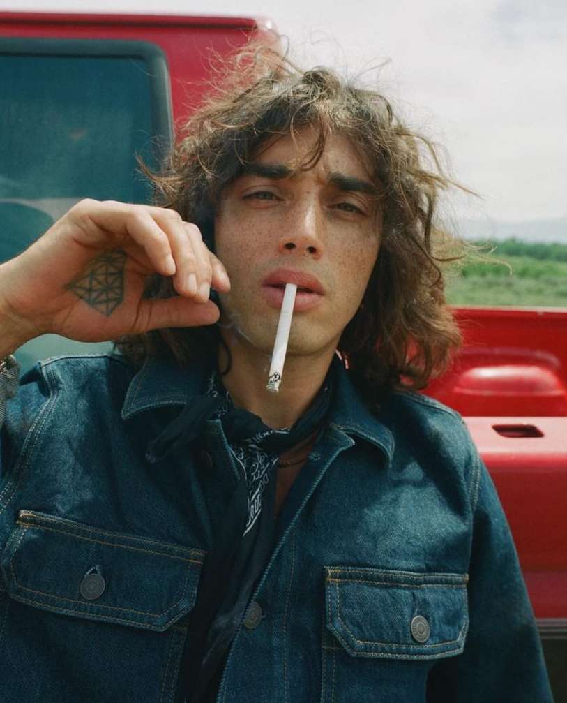 Close-up of a person with wavy brown hair and a denim jacket smoking a cigarette with a red vehicle in the background.