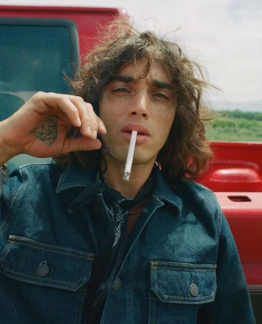 Close-up of a person with wavy brown hair and a denim jacket smoking a cigarette with a red vehicle in the background.