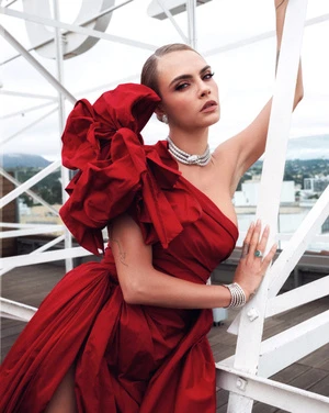 Cara Delevingne poses on a rooftop in a one-shoulder red dress with a large bow, necklace, and bracelets with a cityscape background.