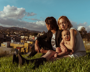 A family sits on a grassy hill overlooking a city, with a mother holding her baby while sitting next to a man in a black suit jacket.