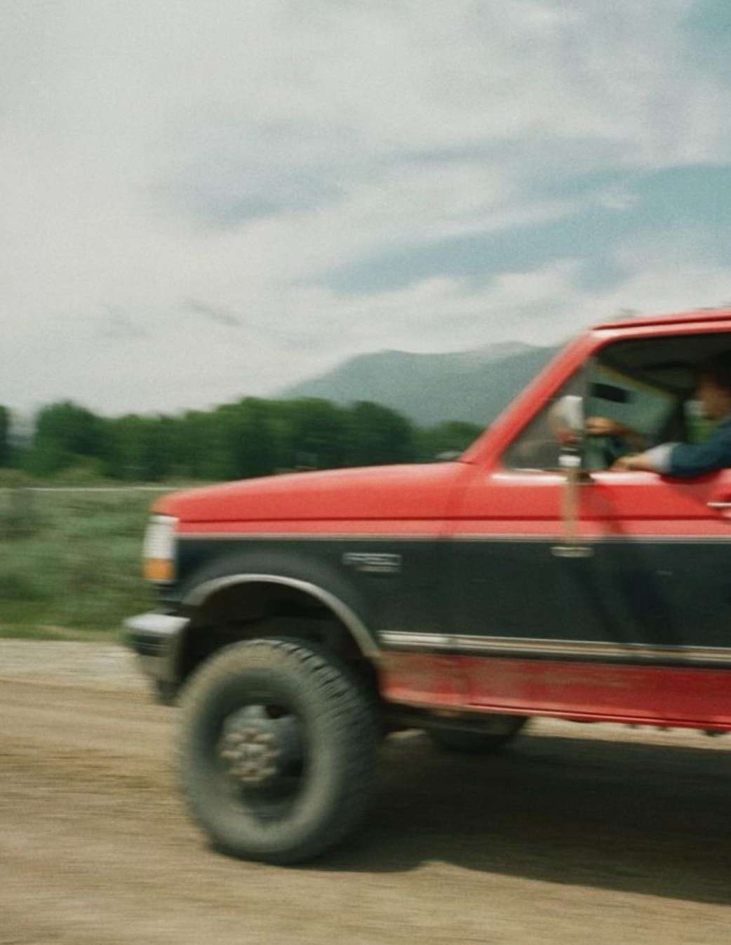 Red and black pickup truck driving on a dirt road with a mountain backdrop and a cloudy sky, creating a sense of motion.
