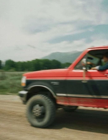 Red and black pickup truck driving on a dirt road with a mountain backdrop and a cloudy sky, creating a sense of motion.